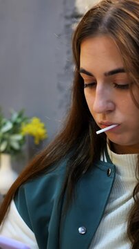 Young caucasian woman with lollipop looking down in cozy cafe, wearing green varsity jacket, scrolling smartphone, soft bokeh background with yellow flowers, pensive expression, warm natural light,