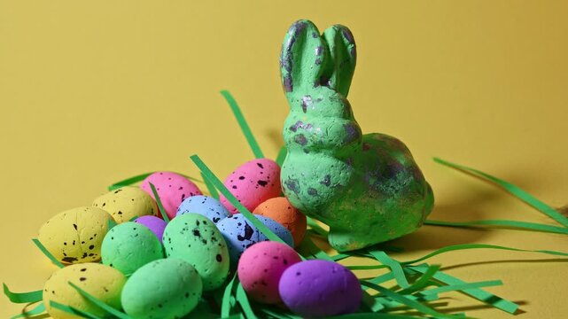 Playful cat paw batting at green glitter Easter bunny and colorful speckled eggs on yellow background. Funny pet behavior during spring holiday