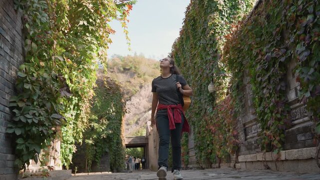 Woman standing in ivy lined passage, curious gaze upward and hands in pockets, red jacket tied at waist, botanist like observation and quiet wonder, cinematic tilt revealing texture and light