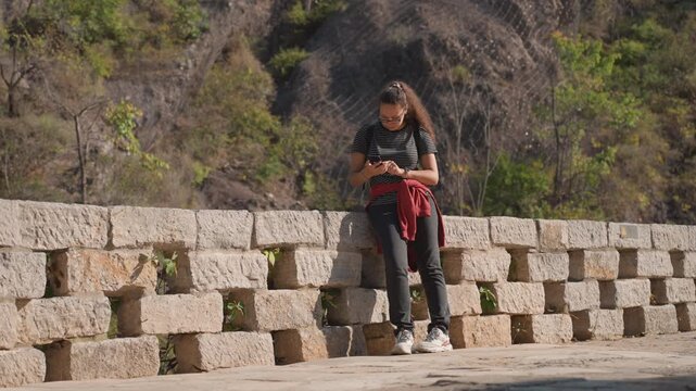 Woman leaning on stone wall texting while pausing on canyon pathway wide shots show casual fullbody pose against textured masonry, sweater and sneakers, checking notifications, adjusting hair