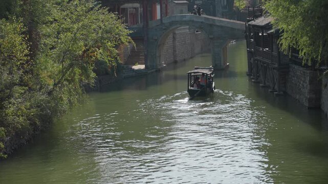 Boat gliding toward arched stone bridge, passengers seated as sunlight shimmers on rippled green water, willow branches and historic houses lining riverside, slow cinematic motion capturing heritage