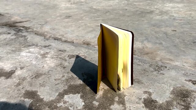 Small book with blank yellow pages standing on a concrete ground with sharp cinematic shadows in high angle shot