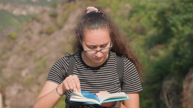 Focused student reading notebook outdoors by hillside, intense concentration with backpack and glasses, flipping pages while studying near rustic village buildings, warm sunlight and candid portrait