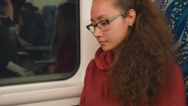 Woman writing notes by train window, red hoodie and glasses, pen on paper at folddown table, concentrated posture with blurred scenery outside, earbuds and timetable on tray, studious atmosphere