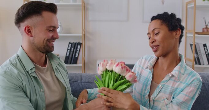 Loving man giving bouquet of flowers to african american woman closing her eyes with her hands, anticipating joyful holiday surprise, celebrate special occasion, romantic gesture expressing affection
