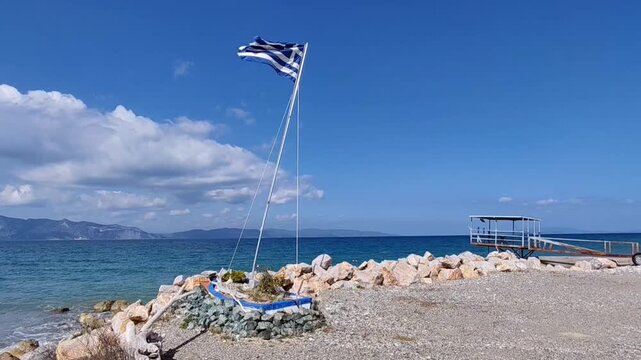 The national flag of Greece waving in the wind on a rocky beach in Evia. A symbolic wooden boat monument against the blue Aegean Sea and sky, capturing the essence of Greek maritime culture