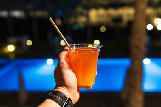Man's hand holding a bright orange cocktail in a plastic cup with paper straw by a blue swimming pool at night