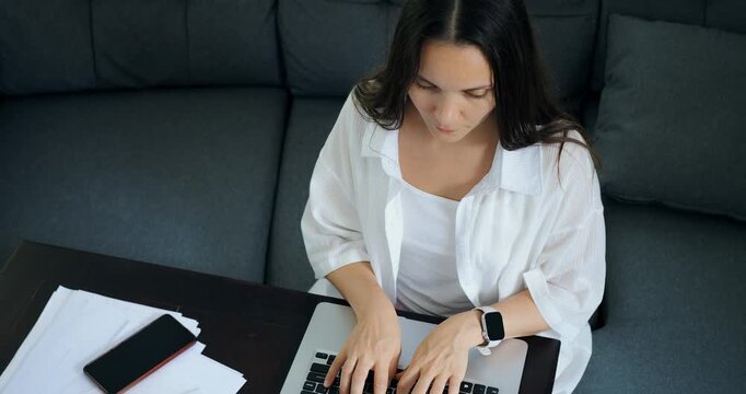 Casual woman working on documents at home. Woman in cozy room reviewing papers and emails on laptop. Relaxed lady sitting on sofa working on documents and checking her smart device