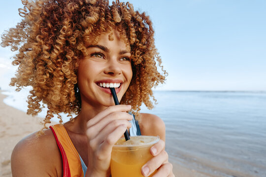 Smiling woman with curly hair drinking orange juice through a straw on the beach. Bright sunny day with clear blue sky and calm sea in the background. Woman lifestyle concept.