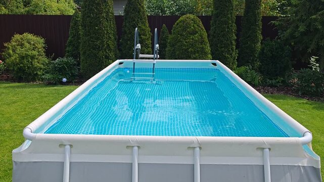 There is a rectangular frame grey PVC swimming pool with clean water, a ladder, filter and pump on the mown lawn. Nearby are flower beds, thujas, lanterns, metal fence and houses. Sunny summer weather