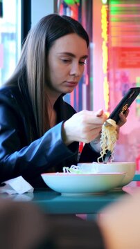 Woman delights in noodles. Casual lady reviews tasty noodles. Vlogger unwinds while sampling spicy hot noodles. Bright neon surroundings complement woman calmly eating her steaming noodles