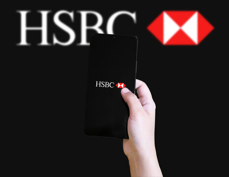 Hand holding smartphone displaying HSBC logo in front of large HSBC logo background illustrative editorial