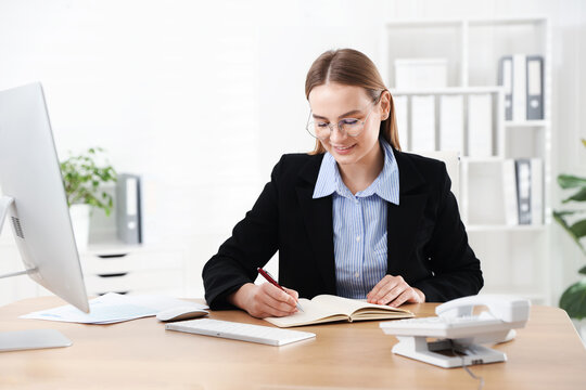 Smiling secretary in suit and glasses taking notes at table in office