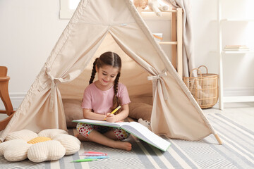 Little girl drawing in sketchbook near toy wigwam at home © New Africa