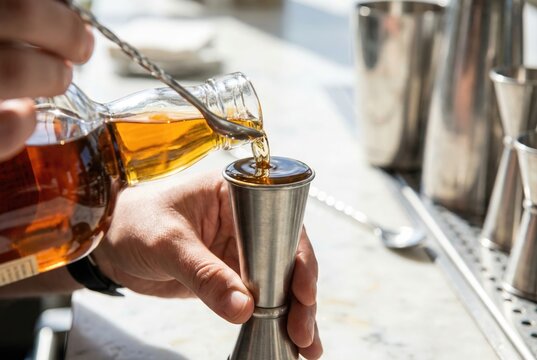 Bartender pouring amber liquor from a bottle into a metal measuring jigger