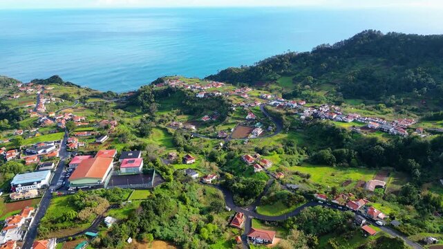 An aerial view reveals the stunning green hills and vibrant Santana villages cascading down to the deep blue Atlantic Ocean on Madeira Island. A winding road connects the charming coastal settlements.