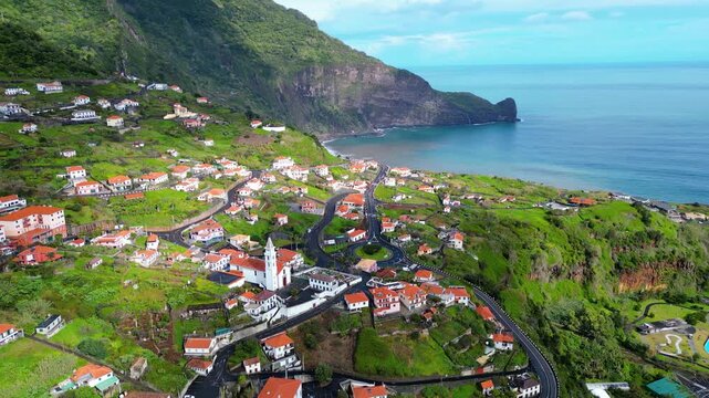 Aerial view of Arco da Faial, Madeira, a charming village nestled on a lush green mountainside. Traditional houses, a church, and winding roads define this picturesque coastal community.