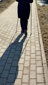 Woman Nordic walking shaded park path, brisk stride with poles and long shadow cast on paving, lea strewn verge, cool crisp light.