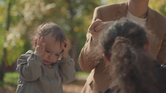 Park emotion scene white parent filming as black caregiver soothes toddler, hands on head, brief fuss then calm, autumn leaves backdrop, attentive exchange, caring family interaction
