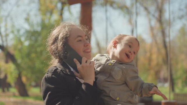 Mother talking on phone while toddler reaches for tree branches candid outdoor family moment in park, sunlight through leaves, attentive parent comforting child, playful exploration and gentle