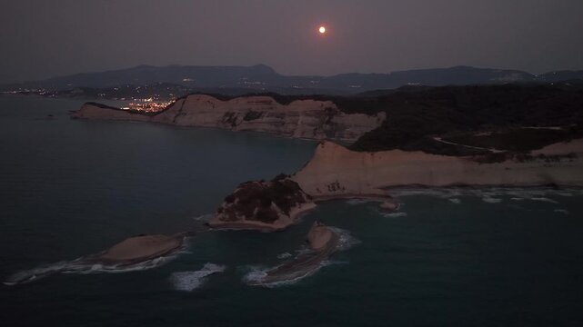 Wide aerial drone shot of a full moon over the dramatic cliffs of Cape Drastis, Corfu, and the glowing lights of a coastal village (like Agios Stefanos) at night in Greece
