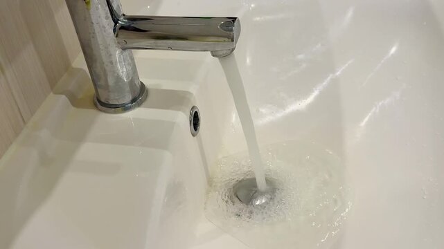 Dirty stainless faucet pouring water into unclean acrylic sink, visible mineral stains and soap scum around drain, slow steady.