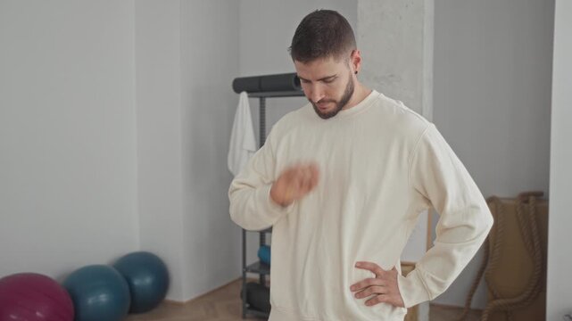 Man touching chin with hand and other hand on hip while checking posture in yoga studio with exercise balls and whiteboard; calm concentration.