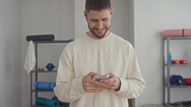 Young hispanic man holding smartphone with bare hands texting amid yoga props and mats in fitness studio; serenity focus wellness.