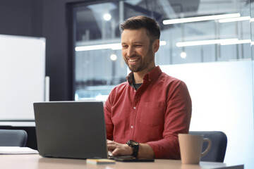 A man sits at a table in an office working on a laptop. He smiles while typing and has a coffee cup beside him. The setting is bright and modern with large windows.