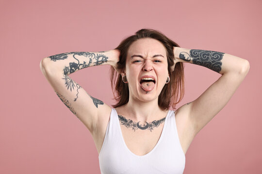 Emotional woman with stylish tattoos and piercings showing tongue on pink background