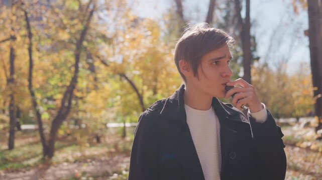 Young white fiance hesitating with vape in autumn park, looking along leaf strewn path, sunlight filtering through trees, worried expression, handheld gesture, decision moment about marriage