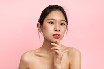 A young woman stands with a thoughtful look on her face. She has one finger on her chin and is facing the camera. The background is a solid pink color, creating a bright atmosphere. © Prostock-studio