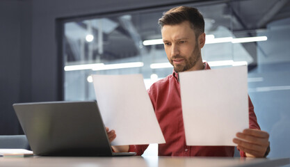 A man sits at a desk in an office looking at two sheets of paper. He appears focused while...