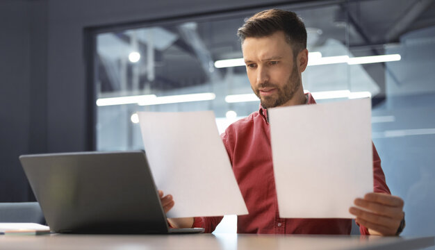 A man sits at a desk in an office looking at two sheets of paper. He appears focused while comparing the documents with a laptop in front of him. Sunlight enters the office space.
