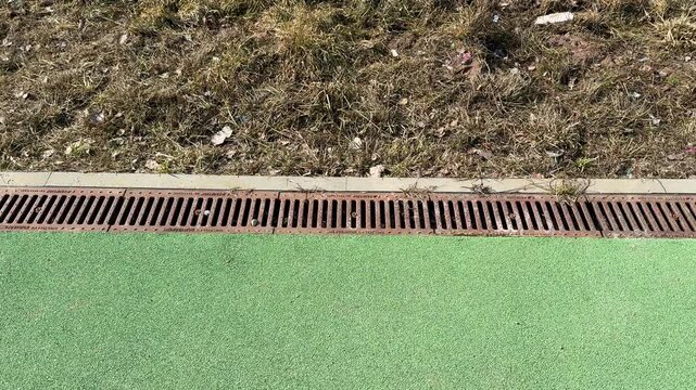 Closeup sports field drainage grate showing rusted iron slots, debris lodged along turf edge, concrete border, textured artificial.
