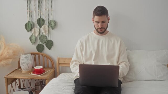Man sips from a red cup held in hand over a laptop while seated on a bed in a bedroom with a wicker bedside table and macrame wall hanging; calm focus.