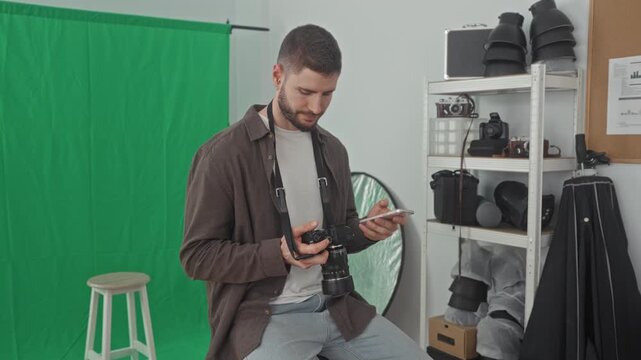 Young man sitting on stool holding dslr camera and checking smartphone near green screen in studio; concentration creativity workflow.