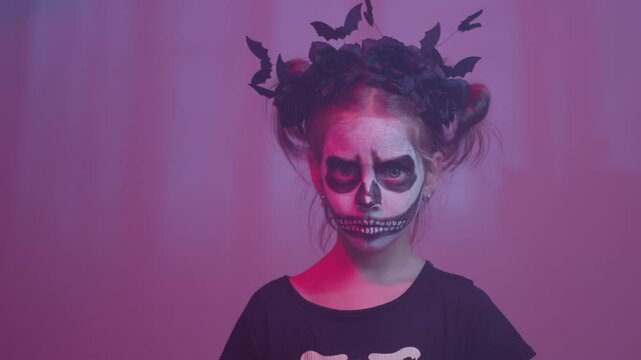 Portrait of little girl wearing skeleton face paint and bat headwear. Child looking at camera with spooky expression creating dark Halloween mood