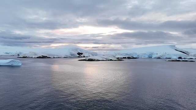 Cinematic aerial moving forward over the calm, dark waters of the Melchior Islands, revealing snow-covered volcanic islets and massive blue-layered ice shelves on the Antarctic Peninsula