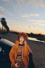 Fototapeta premium Young woman smiling outdoors near car during sunset wearing rainbow sweater and orange jacket, holding smartphone, relaxed lifestyle, casual autumn outfit, natural light