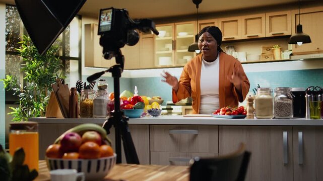 Black woman food vlogger filming culinary content with tripod and camera, behind the scenes of cooking tutorial in home studio kitchen. Influencer ensuring meal prep for online audience.