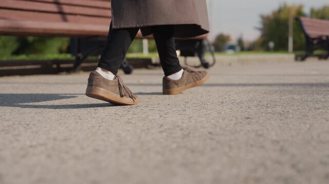 White woman stepping near park bench at morning lowangle closeup on shoes and laces, measured stride and gentle pause, vintage suede footwear, coat hem and shadow play, calm reflective atmosphere,