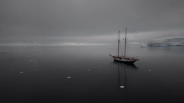 Cinematic aerial orbit of the ice-class schooner Amazone navigating the dark, ice-filled waters of Wilhelmina Bay, set against the massive blue ice cliffs of the Antarctic Peninsula
