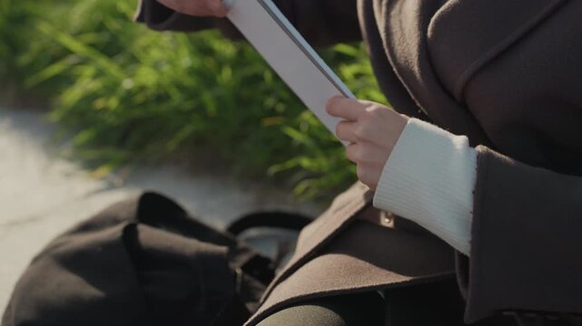 Person opening notebook and writing outdoors, closeup of hands flipping pages and holding pencil, wool coat and backpack at side, sunny urban park setting, focused creative journaling