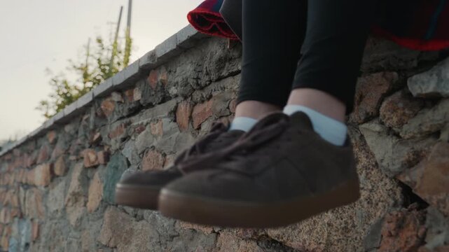 Sunset stone wall closeup shoes dangling. Young student sits on weathered stone ledge, dark sneakers and white socks visible, legs swing slowly against golden backlit sky, urban greenery and soft