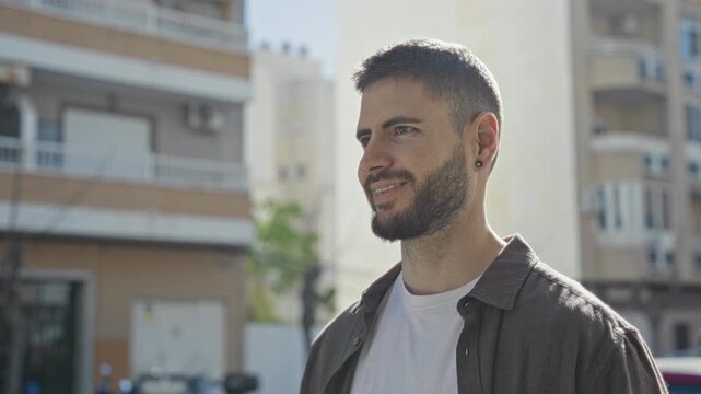 Man smiling showing earring and beard, wearing a casual shirt, standing on a sunlit street by an apartment building and balcony; everyday contentment.
