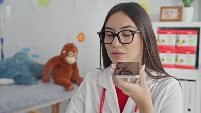 Young woman doctor holds smartphone to mouth while speaking into it in pediatric clinic; compassion care.