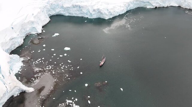 Cinematic descending over the dark, ice-strewn waters of Melchior Harbour toward the expedition schooner Amazone, framed by massive blue ice cliffs in the Antarctic Peninsula