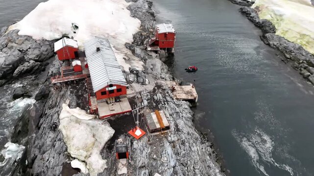 Cinematic aerial orbit of the red wooden buildings and corrugated roofs of Argentina's Melchior Base on Gamma Island, Antarctica as a Zodiac boat with tourists departs the rugged Antarctic shoreline