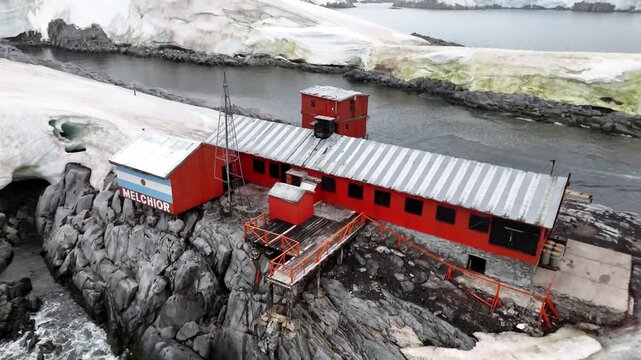 Cinematic aerial raising and tilting down over the abandoned red buildings of Argentina's Melchior Base to reveal the weathered corrugated roofs and rugged basalt cliffs of Gamma Island, Antarctica